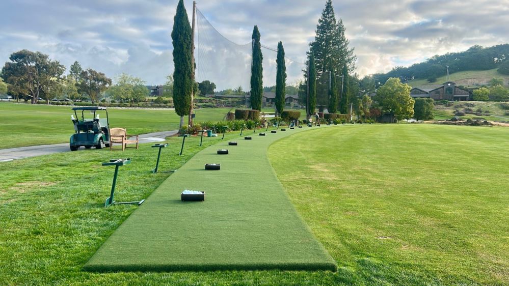 Artificial turf tee lines across a driving range with a golf cart and trees in the background.