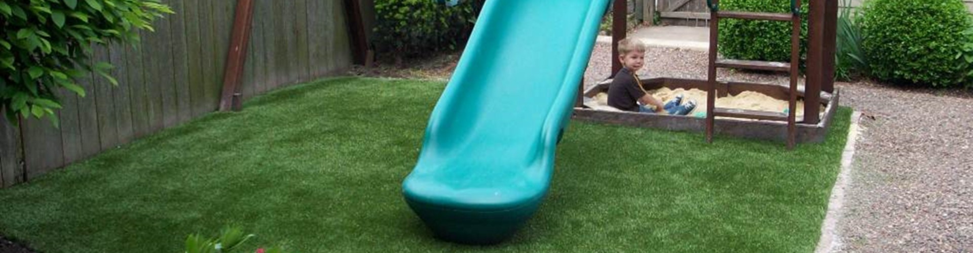 Southwest Greens synthetic grass installation under a backyard playground slide and sandbox with a young child playing safely on artificial turf.