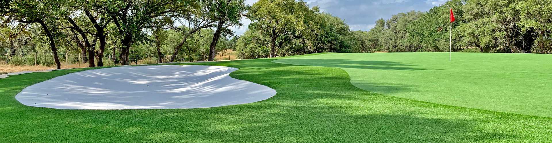 Professionally designed golf green featuring artificial turf with a sand bunker, contoured putting surface, and flagstick set within a scenic landscaped backyard.