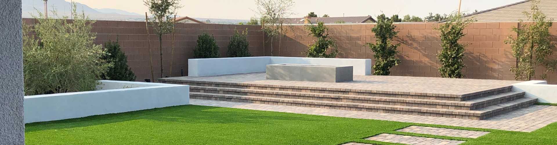 Fake grass installation in a drought-resilient desert backyard with a raised paver patio, modern concrete fire feature, young trees, and stucco block wall landscaping